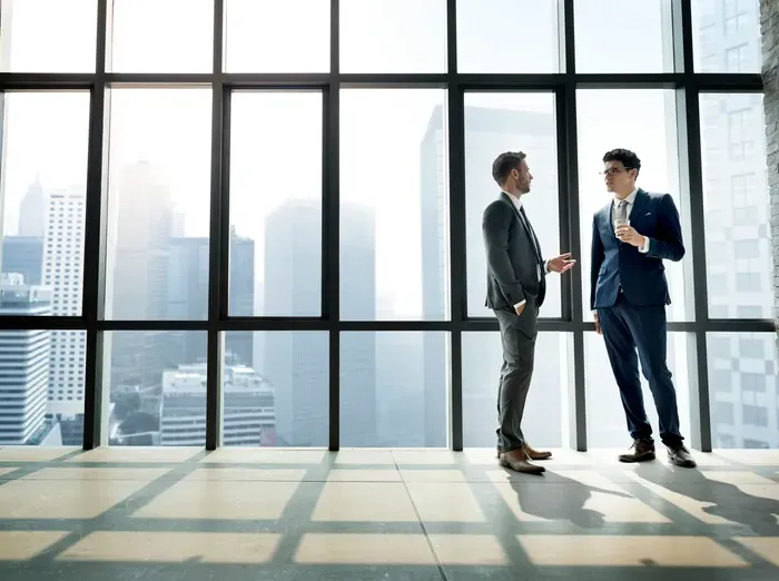 Two professionals in suits chatting by tall windows with city skyline, representing corporate services